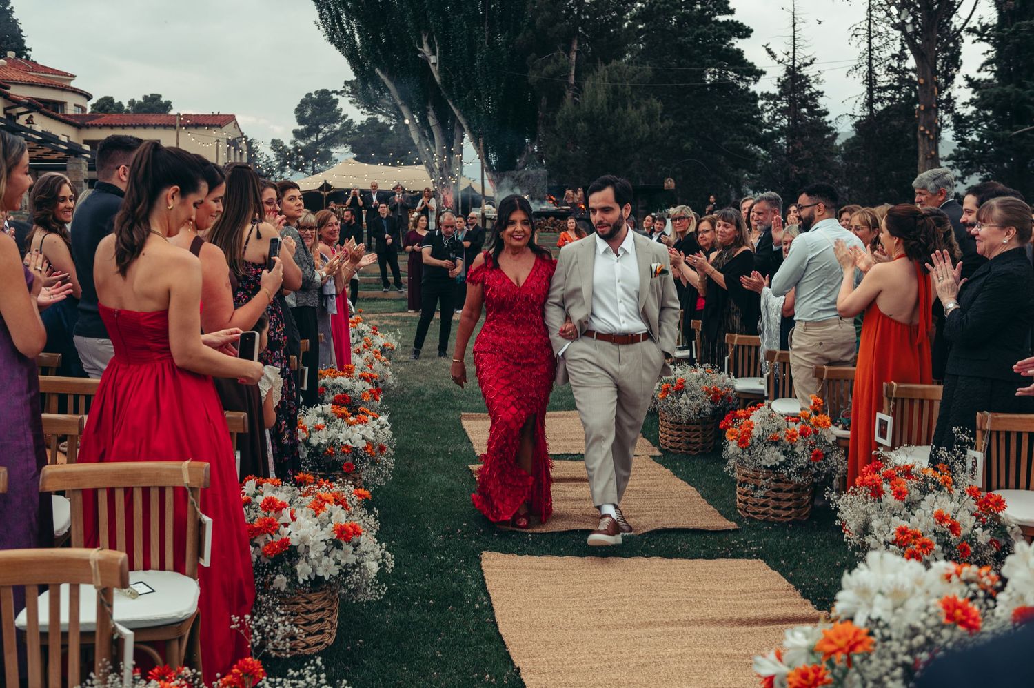 Wedding party in red dresses gathers outdoors for ceremony with floral arrangements and wooden benches.