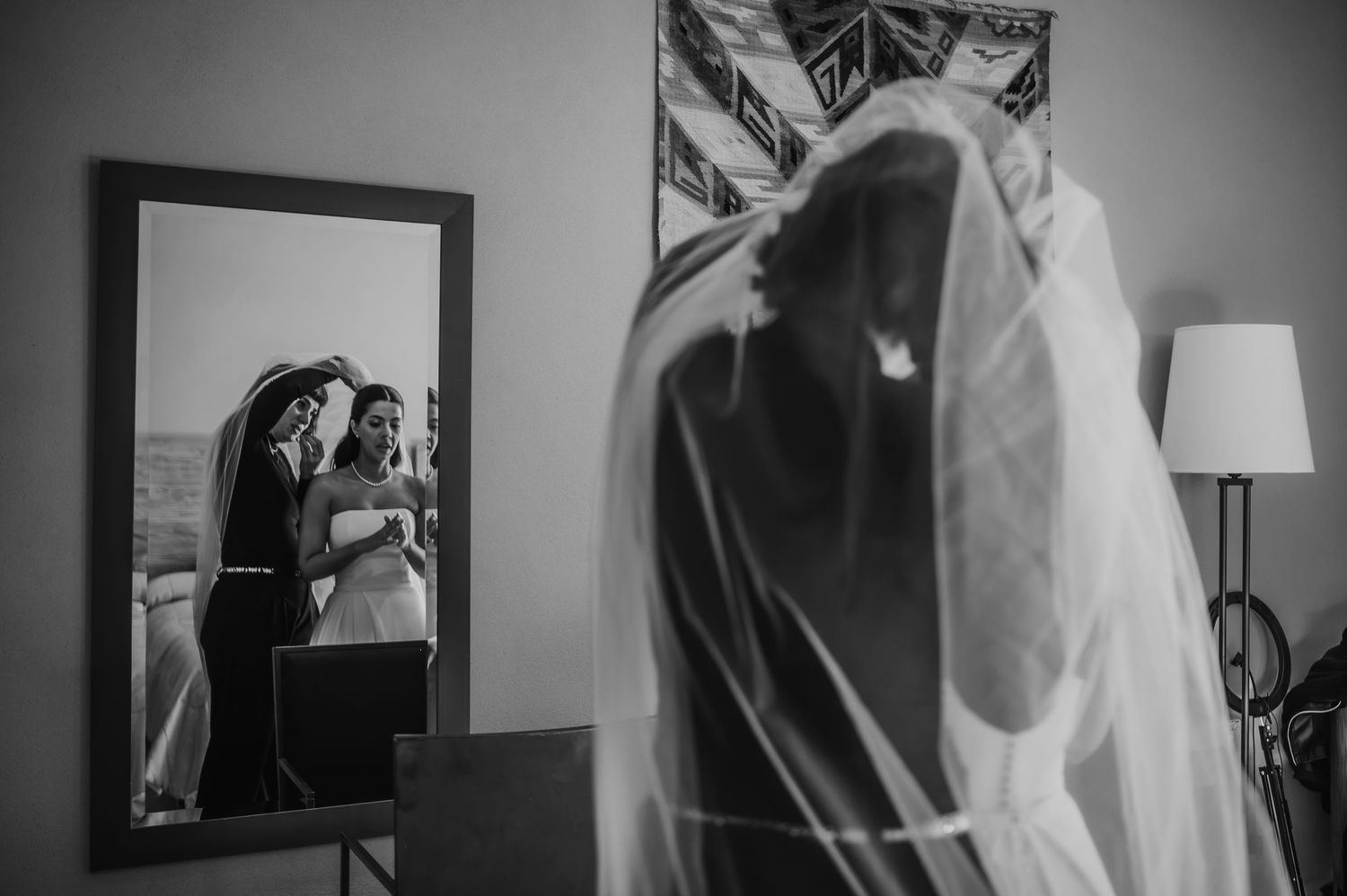 Black and white wedding photo of bride in veil reflected in mirror with bridesmaid during bridal preparations.