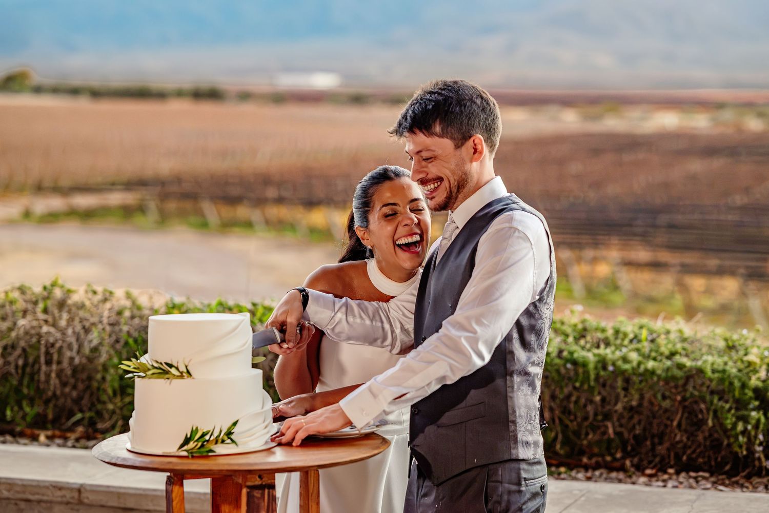 Happy couple cutting their white wedding cake outdoors with scenic vineyard and mountains in background.