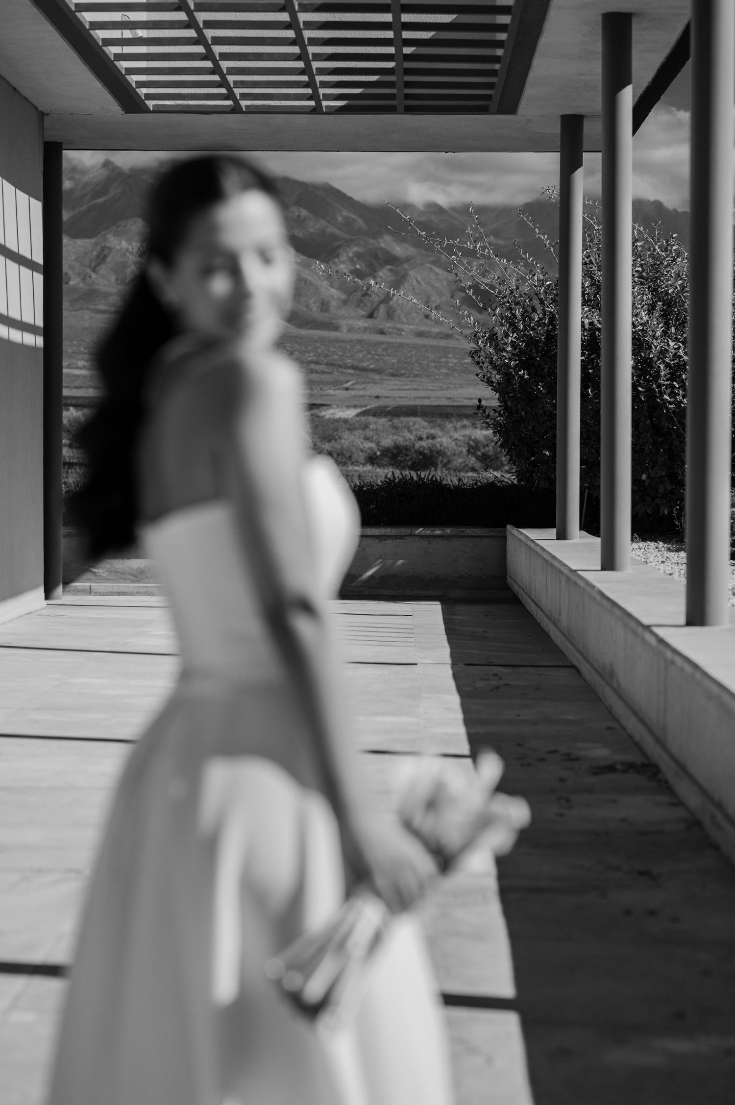 Black and white photo of a pregnant woman in a white dress on a covered patio with mountain views.
