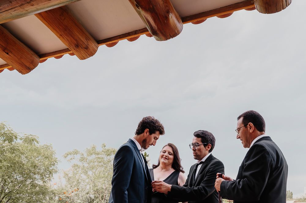 Groomsmen in dark suits gather outdoors under wooden beams during wedding ceremony. wedding in mendoza