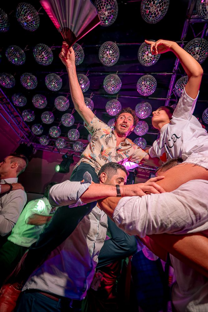Energetic wedding party members performing dynamic dance moves under purple disco lighting and mirror balls.