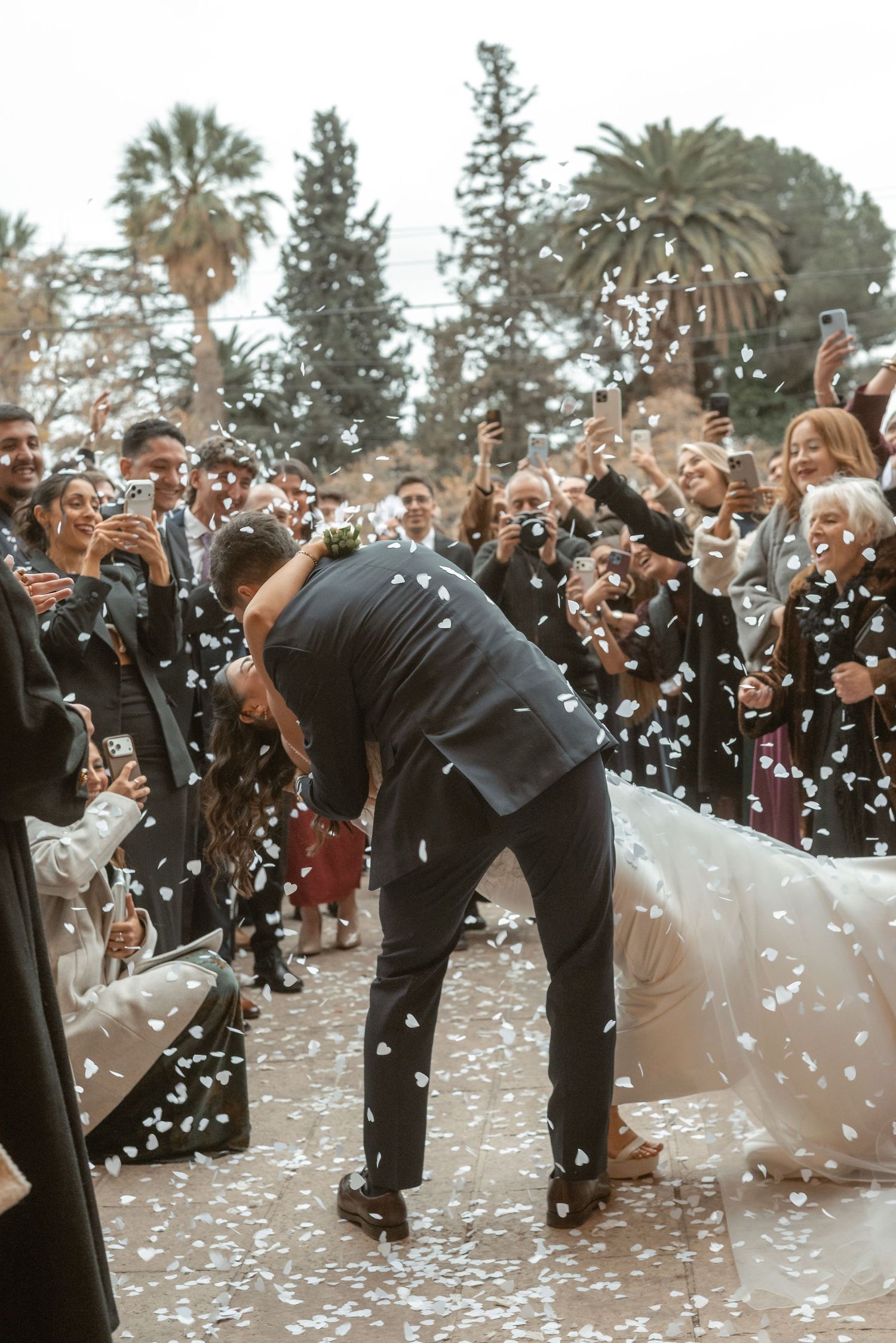Bride and groom share a kiss as confetti falls while wedding guests celebrate and photograph the joyful moment outdoors.