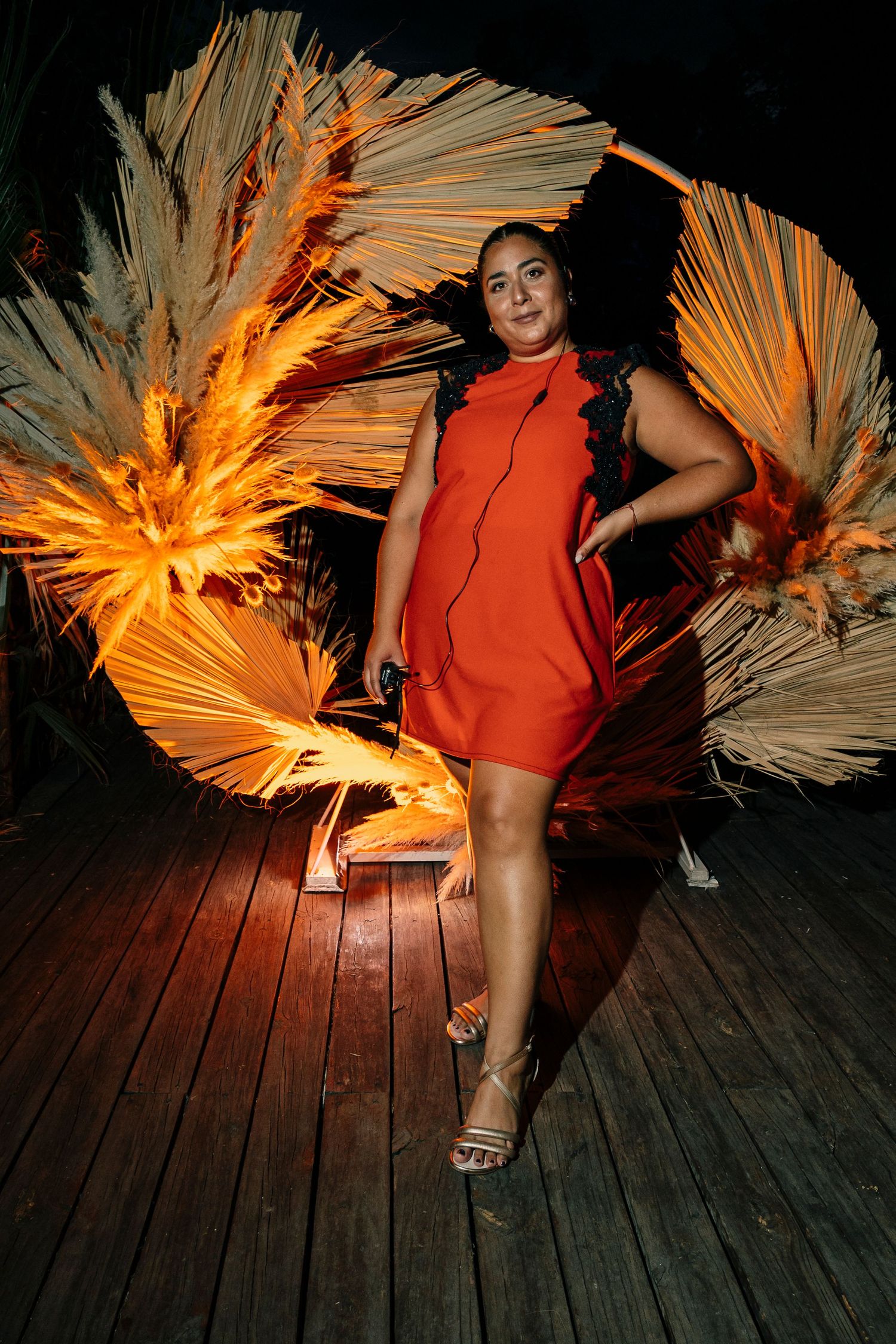 A person in a vibrant orange mini dress poses against a dramatic backdrop of illuminated dried palm fronds on a wooden deck.