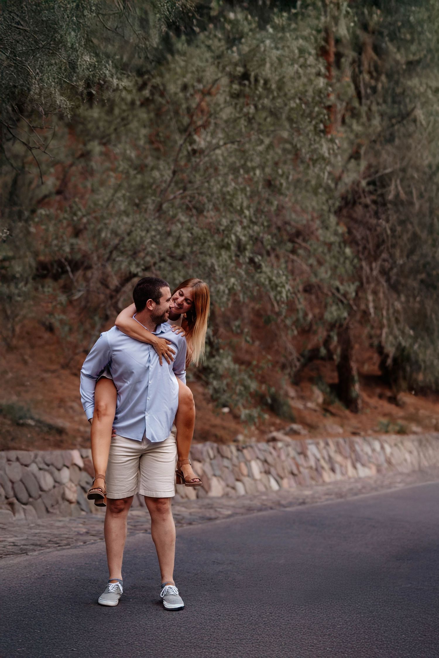A couple embraces on a quiet road surrounded by trees and stone walls at dusk.