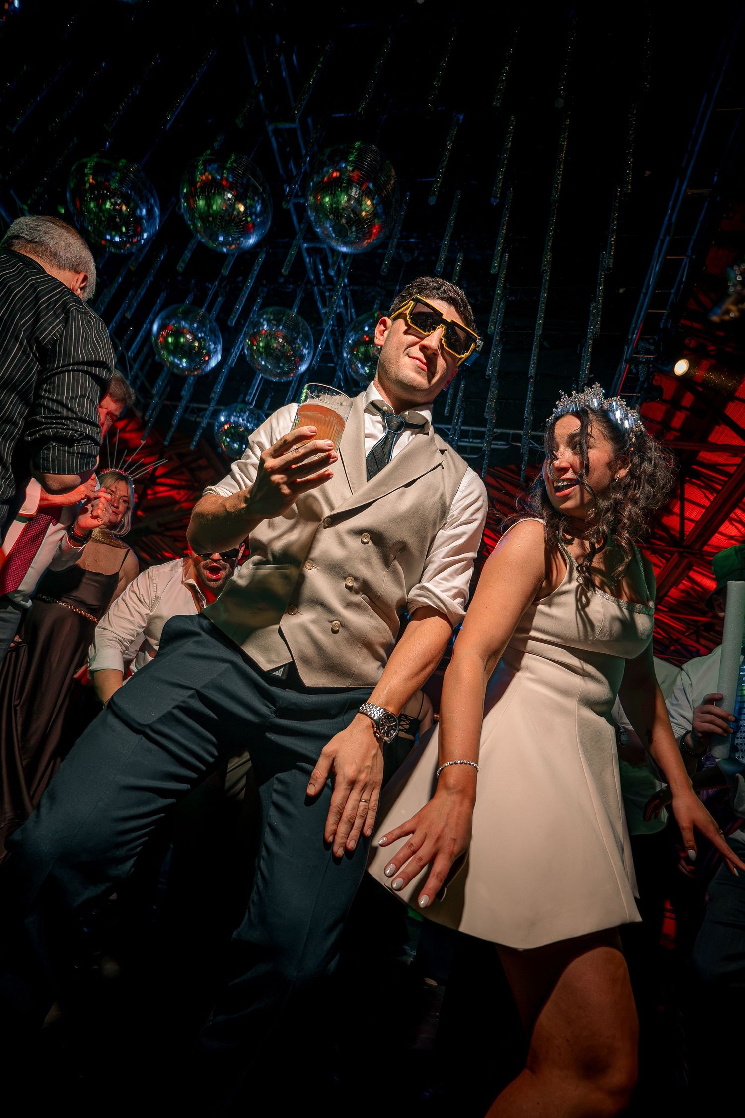 Couple dancing at a festive party with disco balls overhead, man in vest and sunglasses holding drink, woman in white dress.