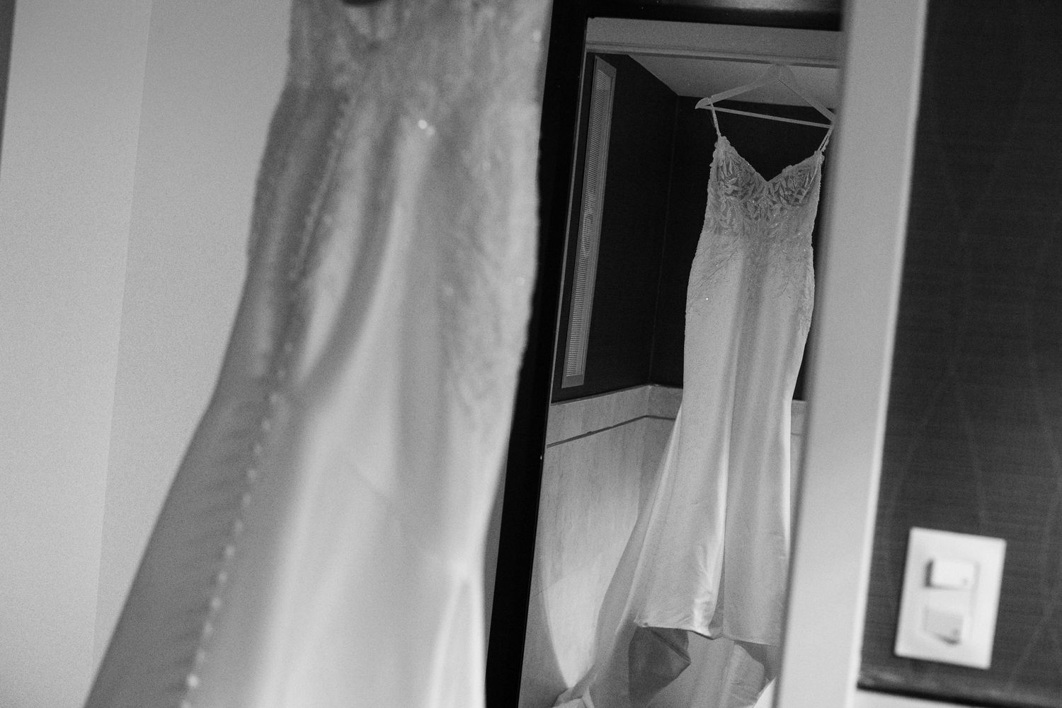Black and white photo of two elegant wedding dresses hanging, one lace and one sleek, displayed in a bridal suite.