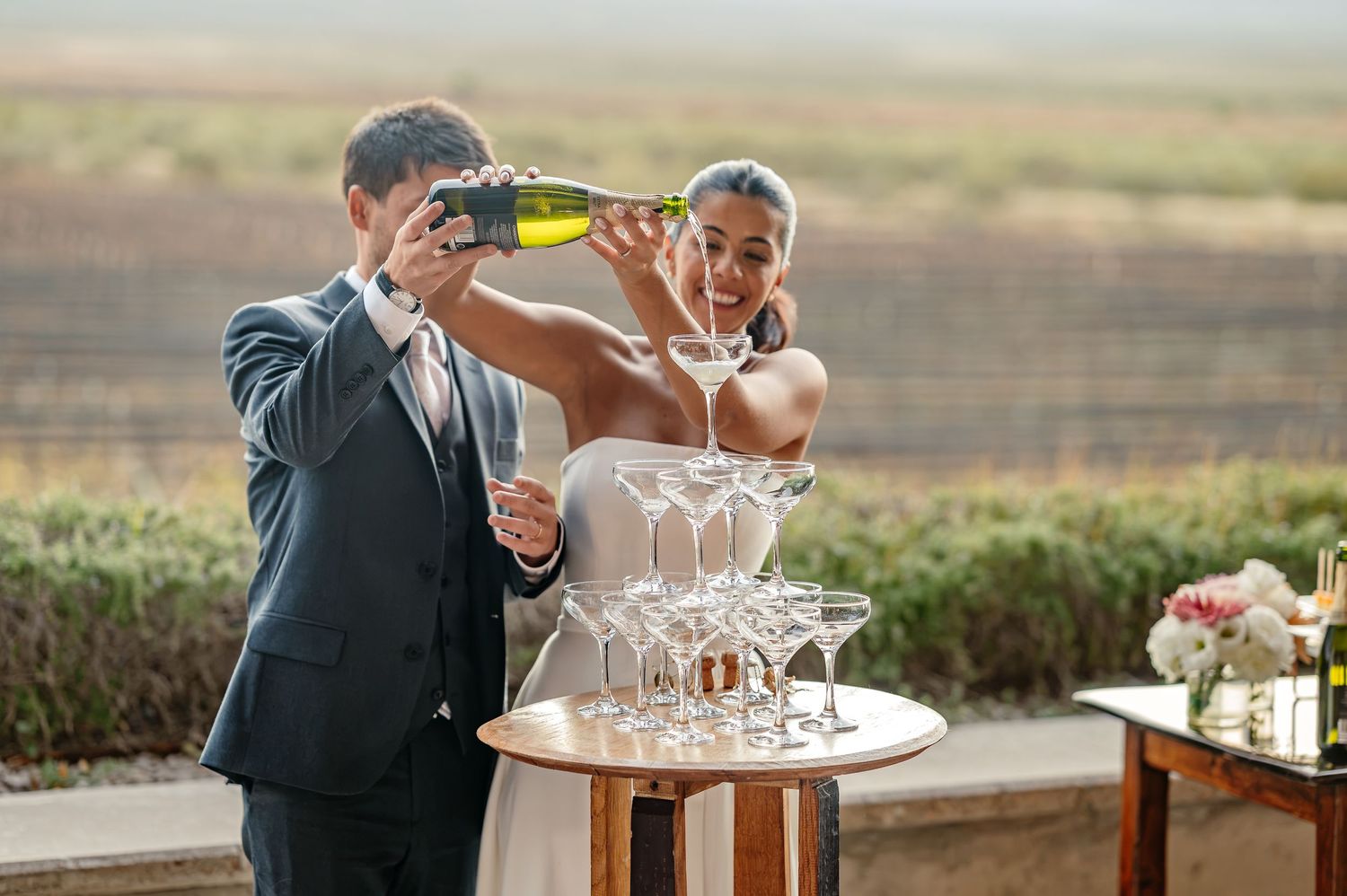 Bride and groom pouring champagne tower at outdoor wedding reception with scenic vineyard backdrop.