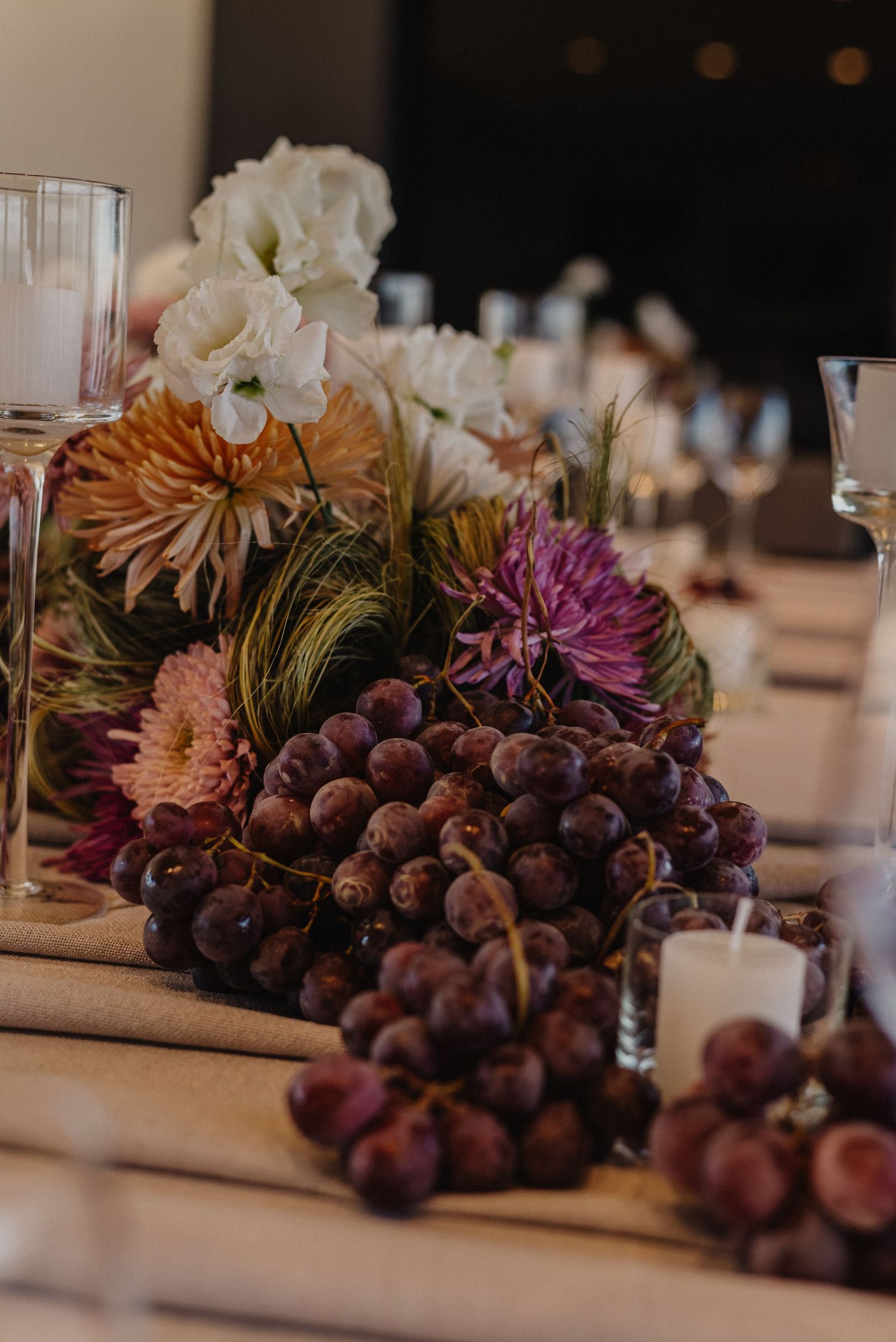 Elegant table centerpiece with purple grapes, white and mauve flowers, and candles for a wedding reception.