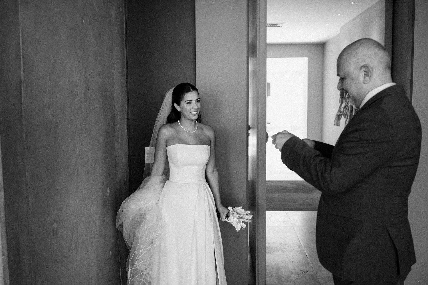 Black and white photo of a bride in a white gown and veil receiving a gift from a bald man in a suit.