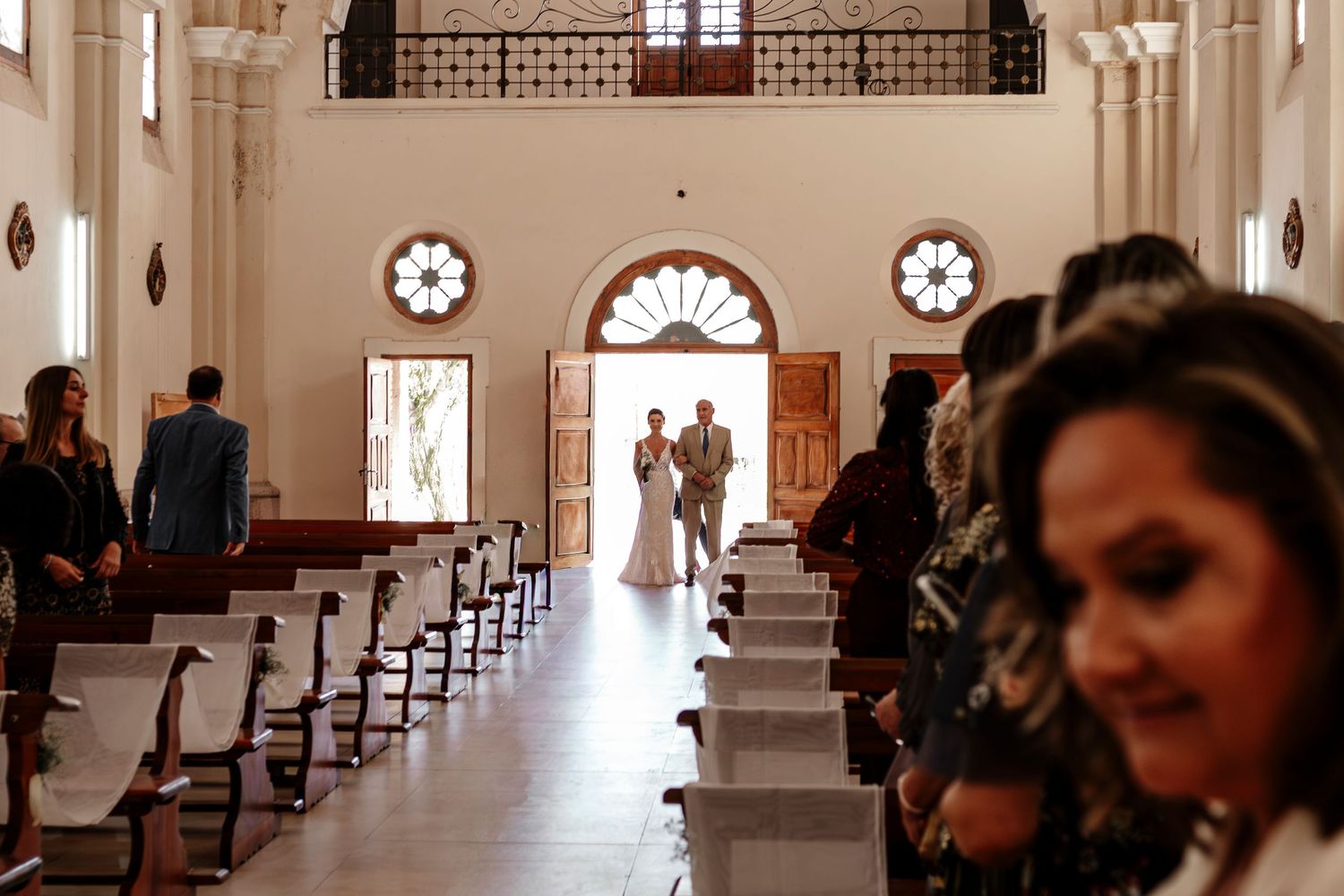 Guests gather in an elegant white church interior with arched windows and ornate architectural details during a ceremony.