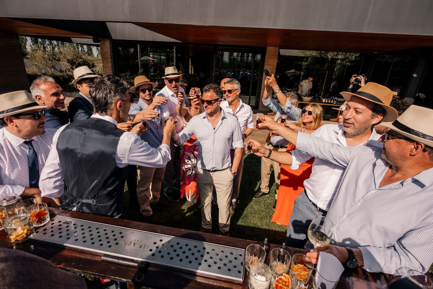 People enjoying drinks and socializing at an outdoor garden party with a bar and lush greenery in the background.