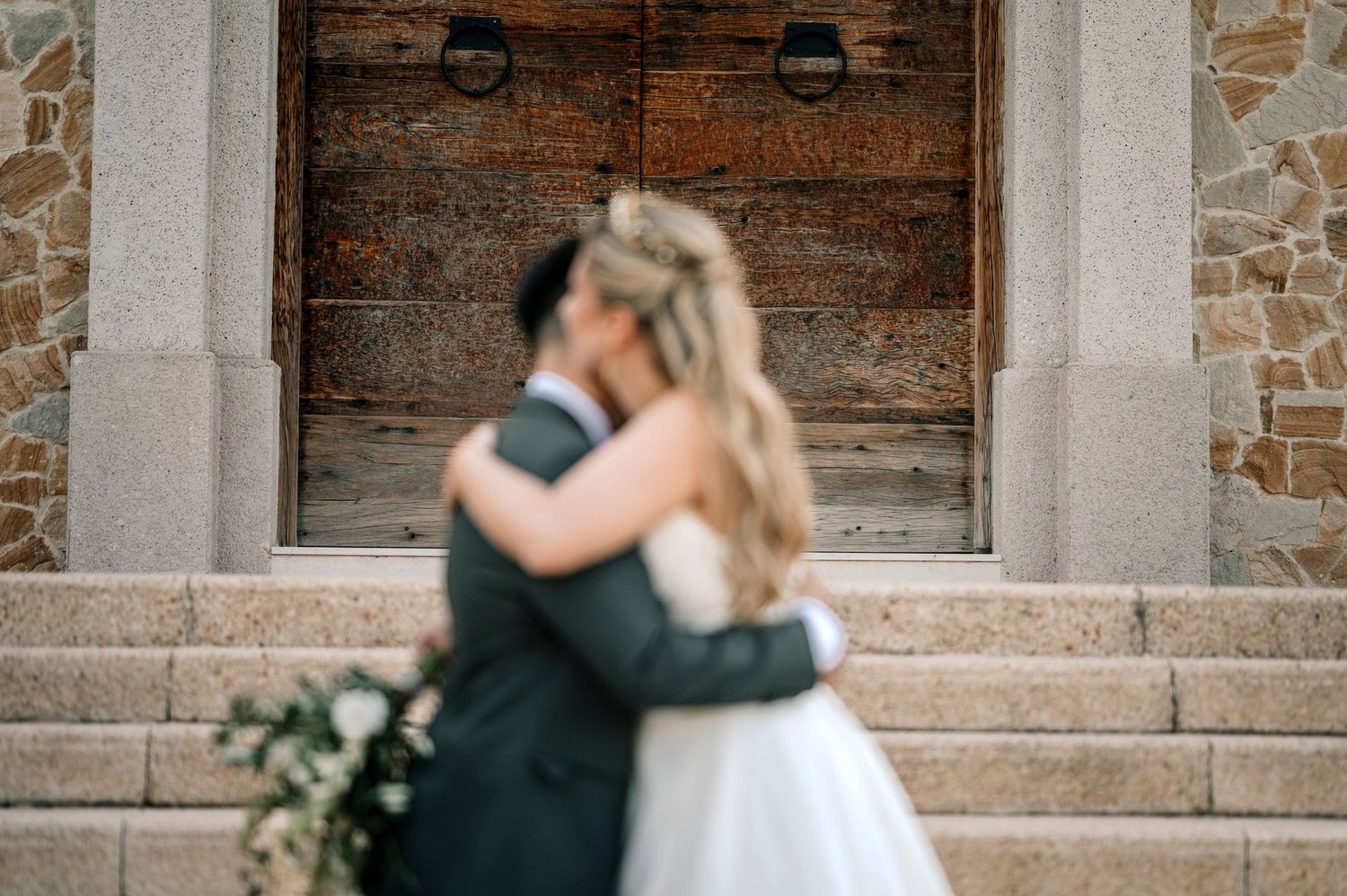 A couple in formal wedding attire shares a romantic kiss against a rustic brick wall backdrop.