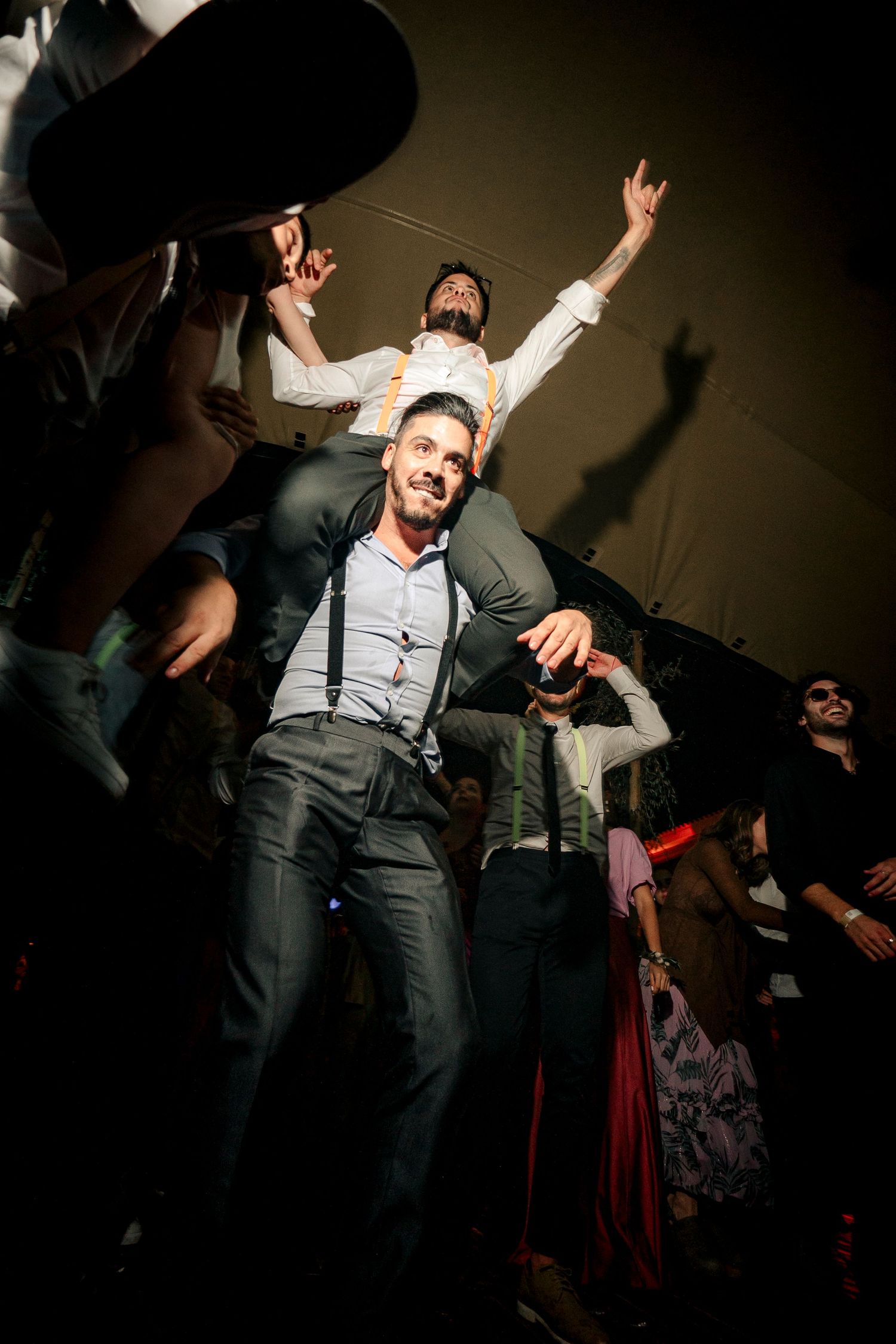 A person in formal attire dances energetically at a party with raised arms and suspenders showing excitement.