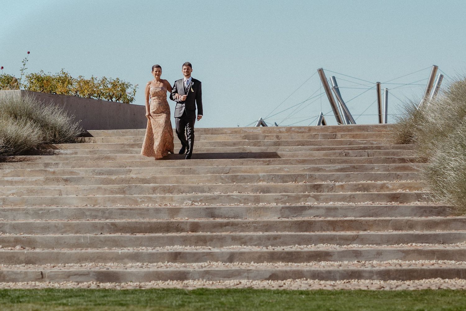 Elegant couple in formal attire walking down outdoor stone steps at a scenic venue on a clear day.