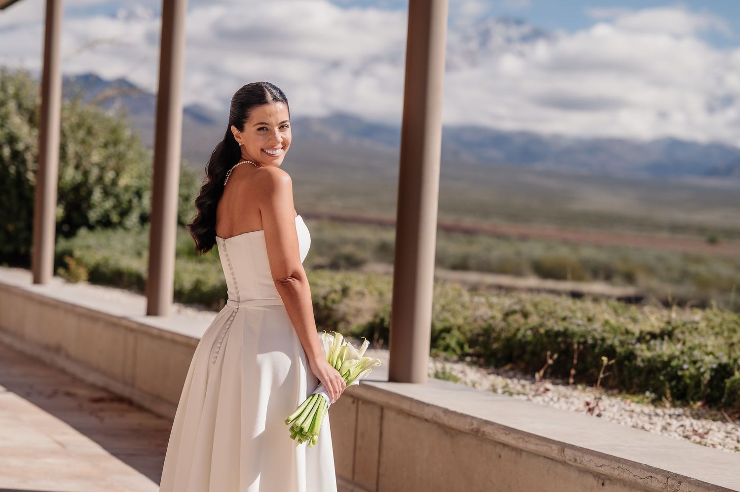 Smiling bride in strapless white gown holds green bouquet on scenic outdoor venue with mountain views.
