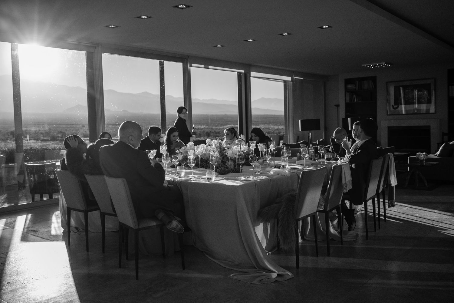 Black and white photo of elegant dinner party at round table with panoramic city views through floor-to-ceiling windows at sunset.