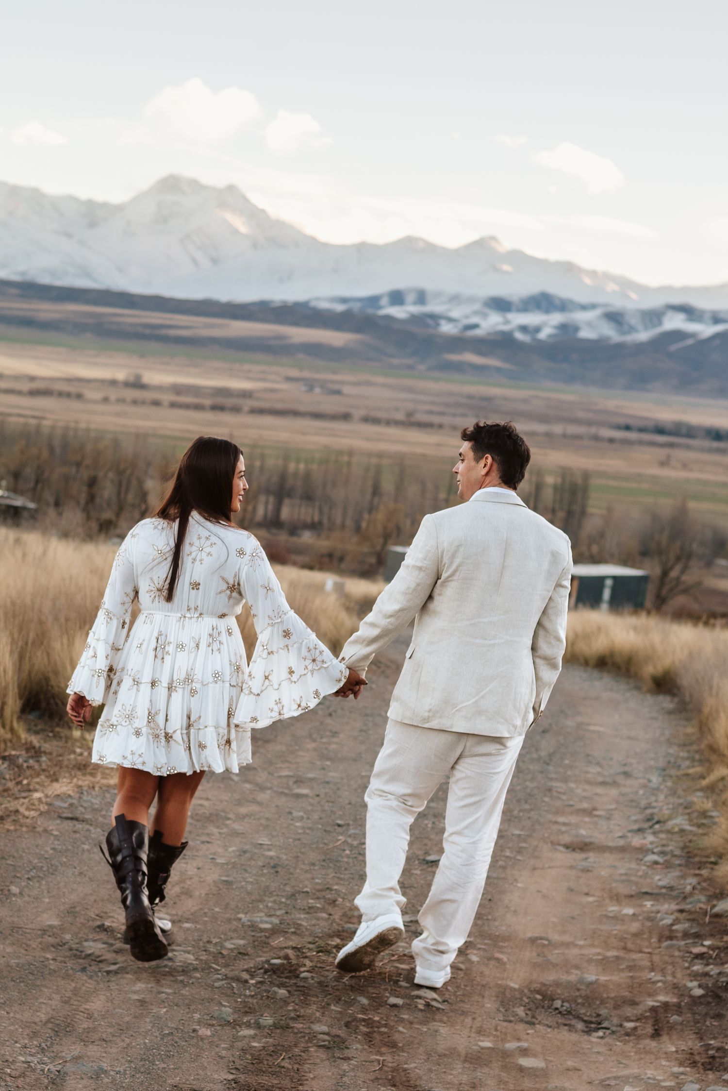 Una pareja camina de la mano por un sendero de tierra con montañas nevadas de fondo; pre boda en mendoza