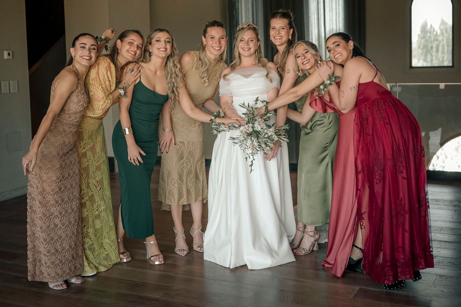 A wedding party poses together in a formal indoor setting wearing burgundy, green and neutral colored dresses.