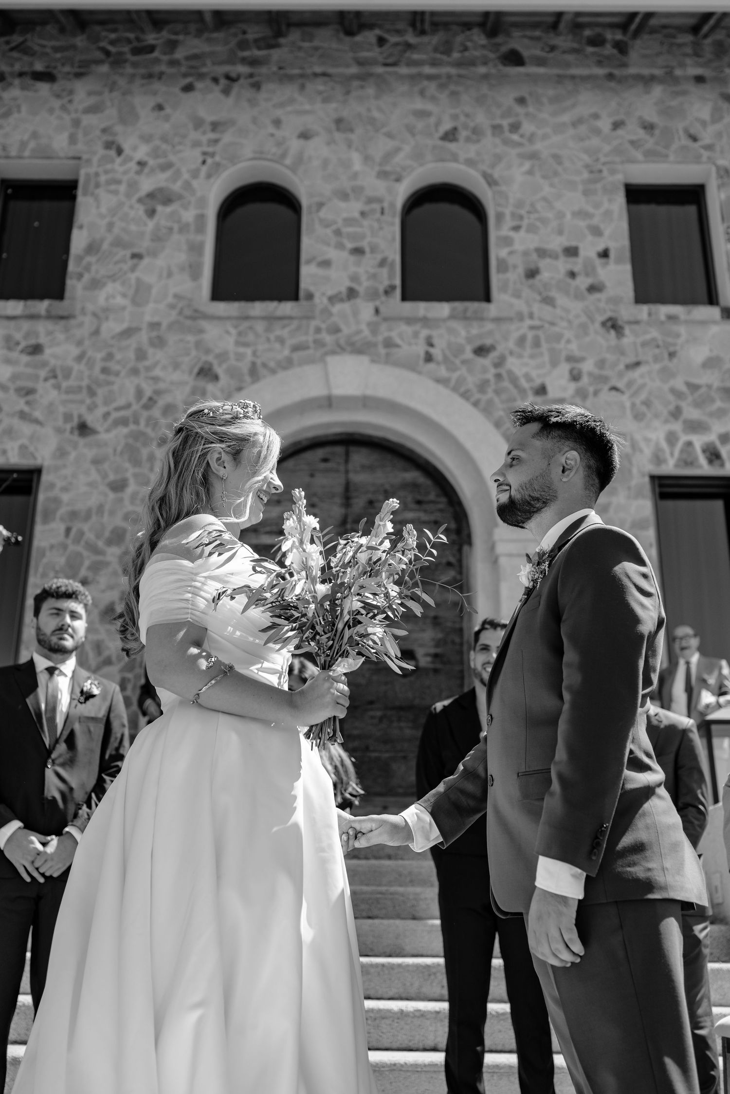 Wedding party in formal attire stands in front of stone building with arched windows during outdoor ceremony.