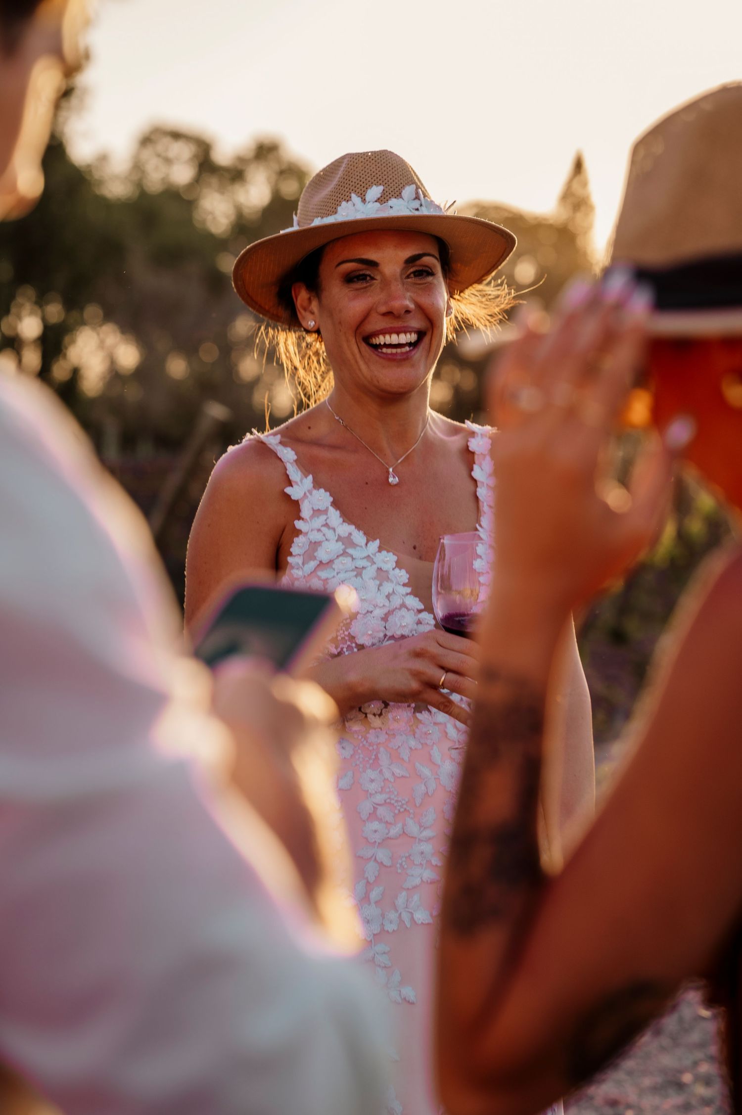 A person in a sun hat and white lace dress laughs joyfully during an outdoor sunset gathering.