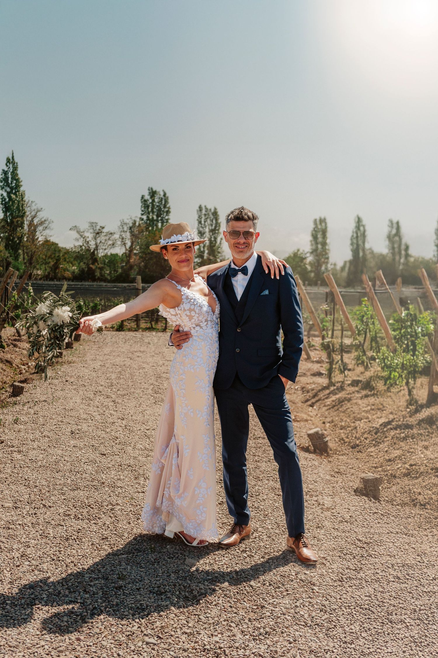 A couple in wedding attire poses together in a rustic vineyard setting on a sunny afternoon.