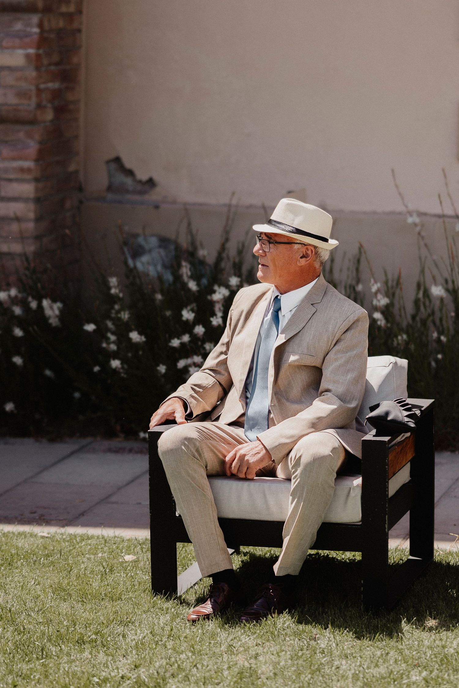 A person in a light colored suit and hat sits relaxed on a bench in a sunny outdoor setting.