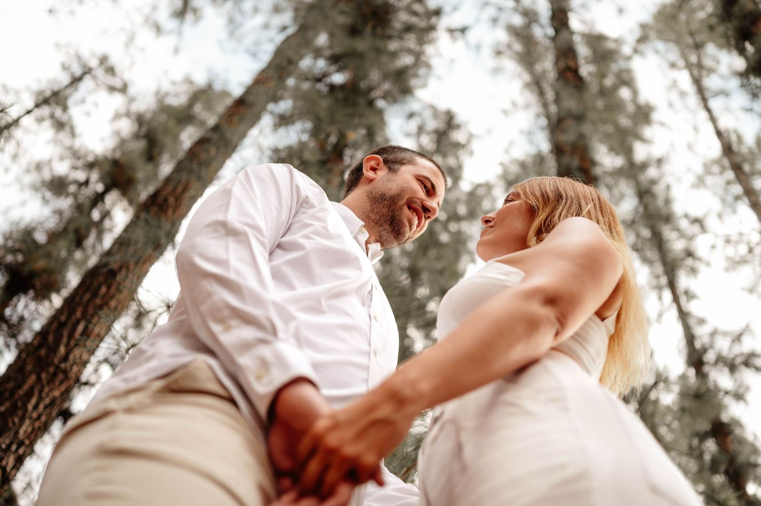A romantic couple in wedding attire share a joyful moment together beneath tall pine trees during an outdoor celebration. preboda in mendoza