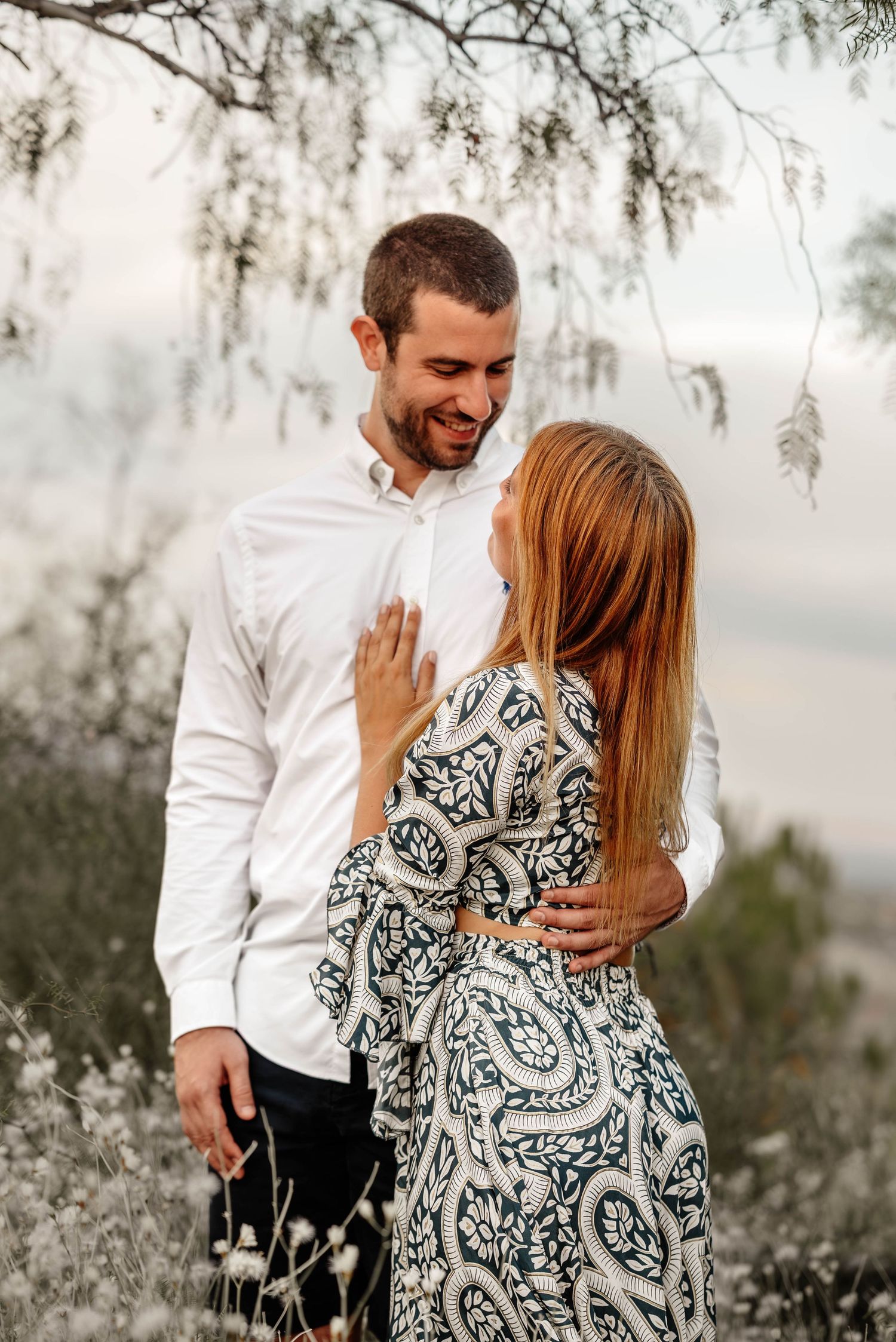 A romantic couple embraces under willow branches in soft lighting, with her wearing a patterned dress and him in white.