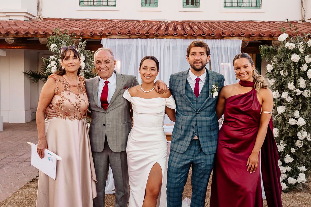 Wedding party in coordinated burgundy, gray and white attire poses together in front of a light colored building. wedding in mendoza