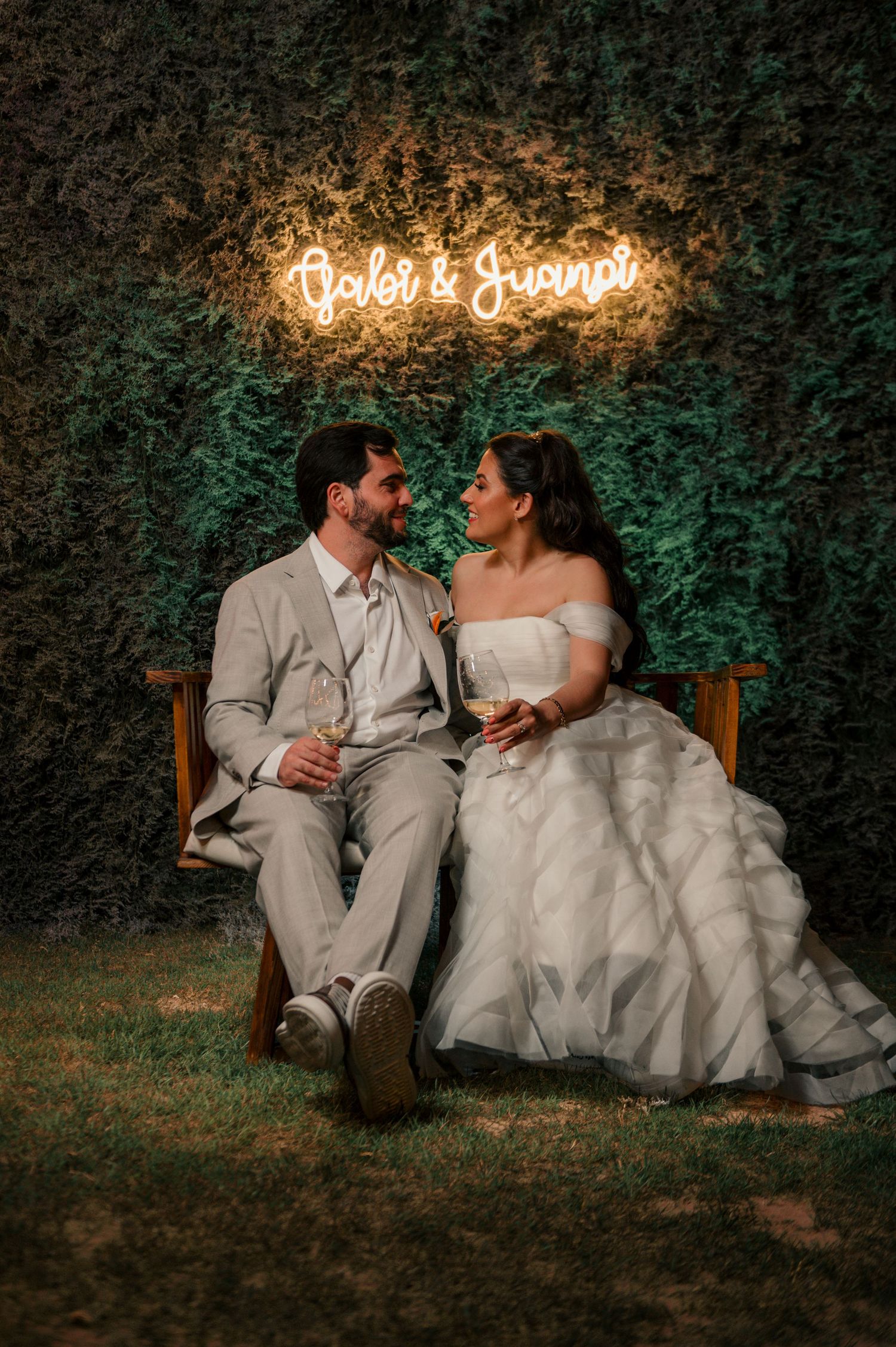 A romantic wedding couple sits together on rustic wooden chairs against a green hedge backdrop with glowing neon signage.