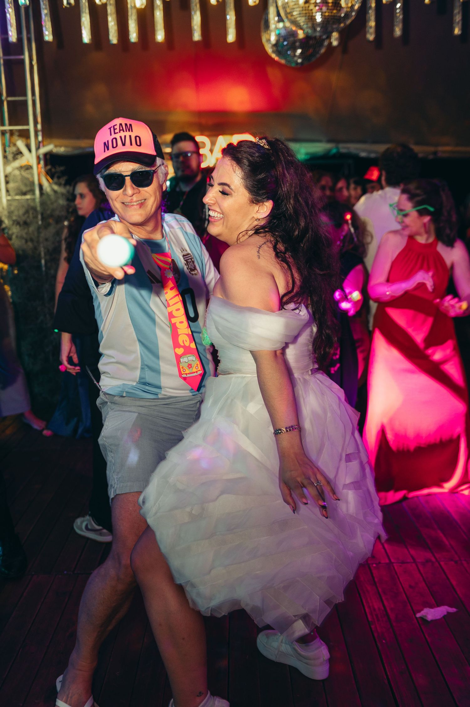 Wedding couple dances energetically on a dark dance floor with colorful pink lighting and disco ball overhead. fotografos de boda en mendoza