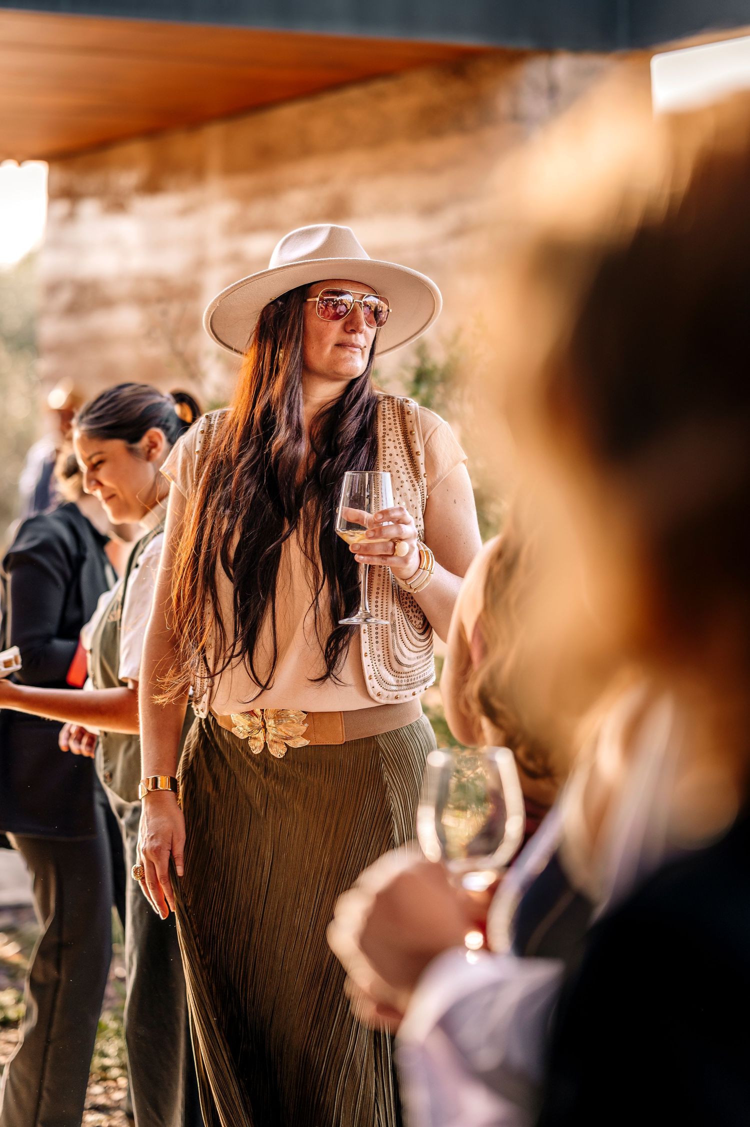 Woman in a wide-brimmed hat and flowing dress mingles at an outdoor social gathering.