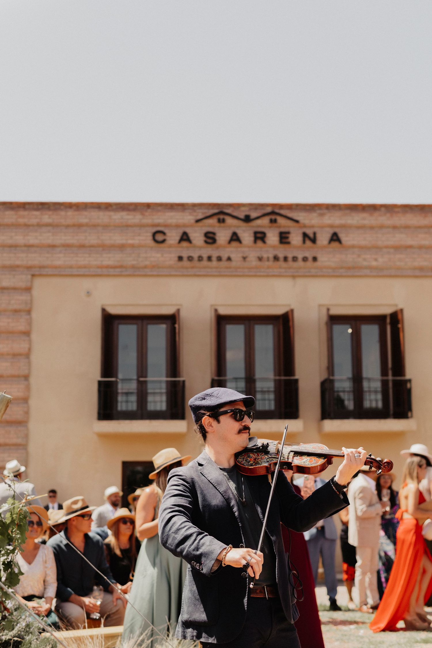Musicians in sunglasses and formal attire perform on violins at an outdoor event with guests in the background.