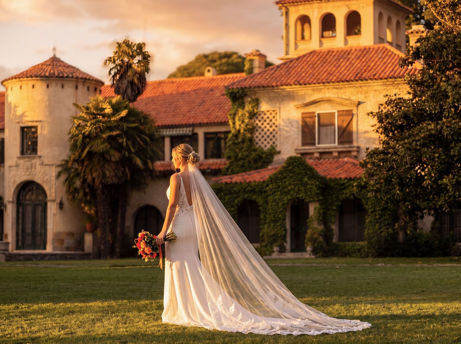 Bride in white gown with long veil stands on green lawn before ivy-covered Spanish villa at golden sunset.