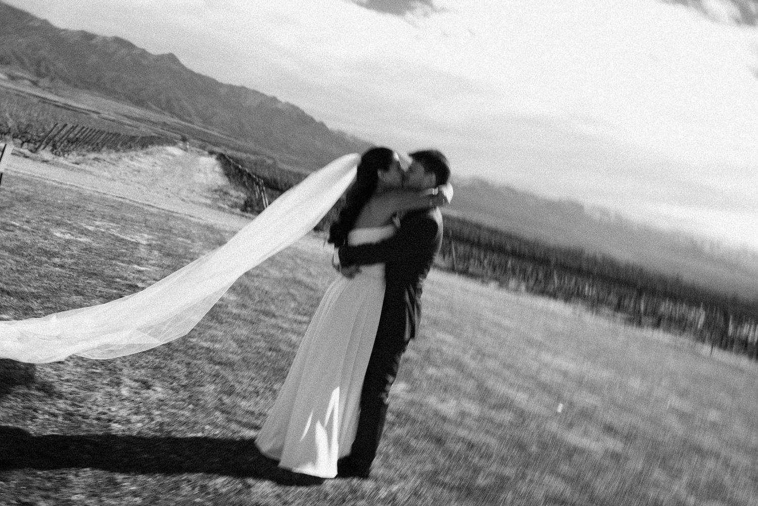 Black and white wedding photo of couple kissing in field, bride's veil flowing in wind with mountain backdrop.