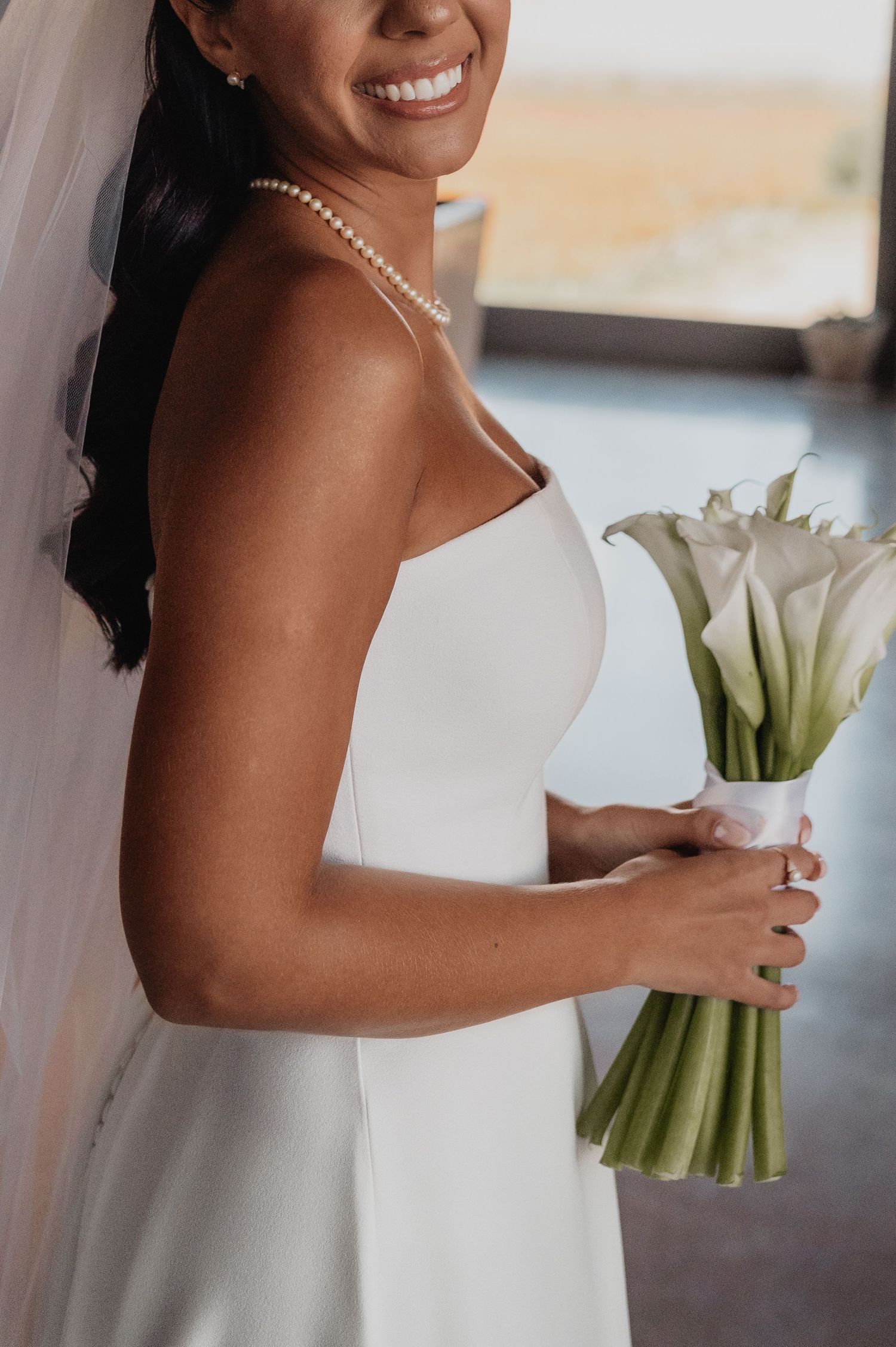 Smiling bride in white strapless gown holding calla lily bouquet, wearing pearl necklace and veil. fotógrafos de boda Mendoza