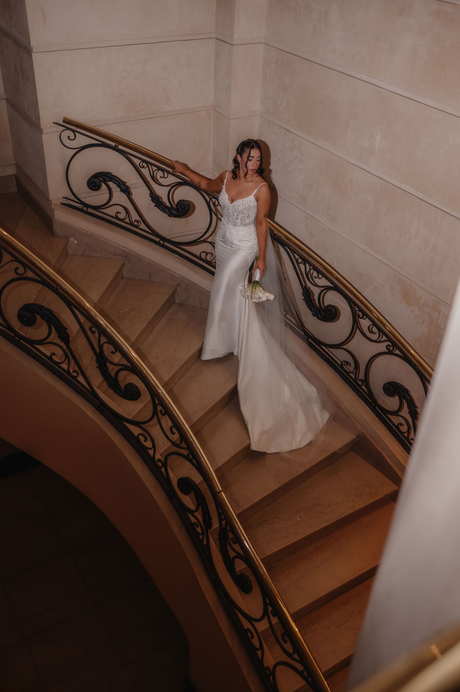 Novia con vestido de novia blanco sosteniendo un ramo en una elegante escalera curva con barandillas de hierro ornamentadas.