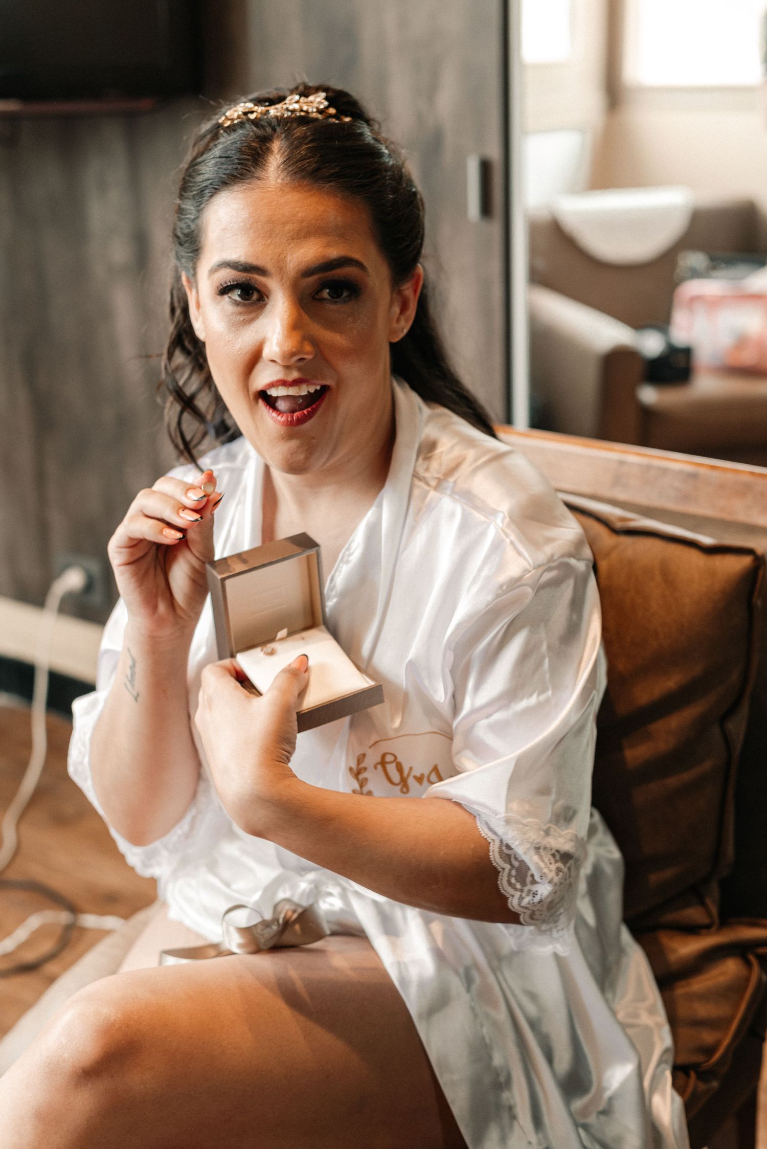 A person in a white silk robe holds a small jewelry box while getting ready on a brown leather couch.