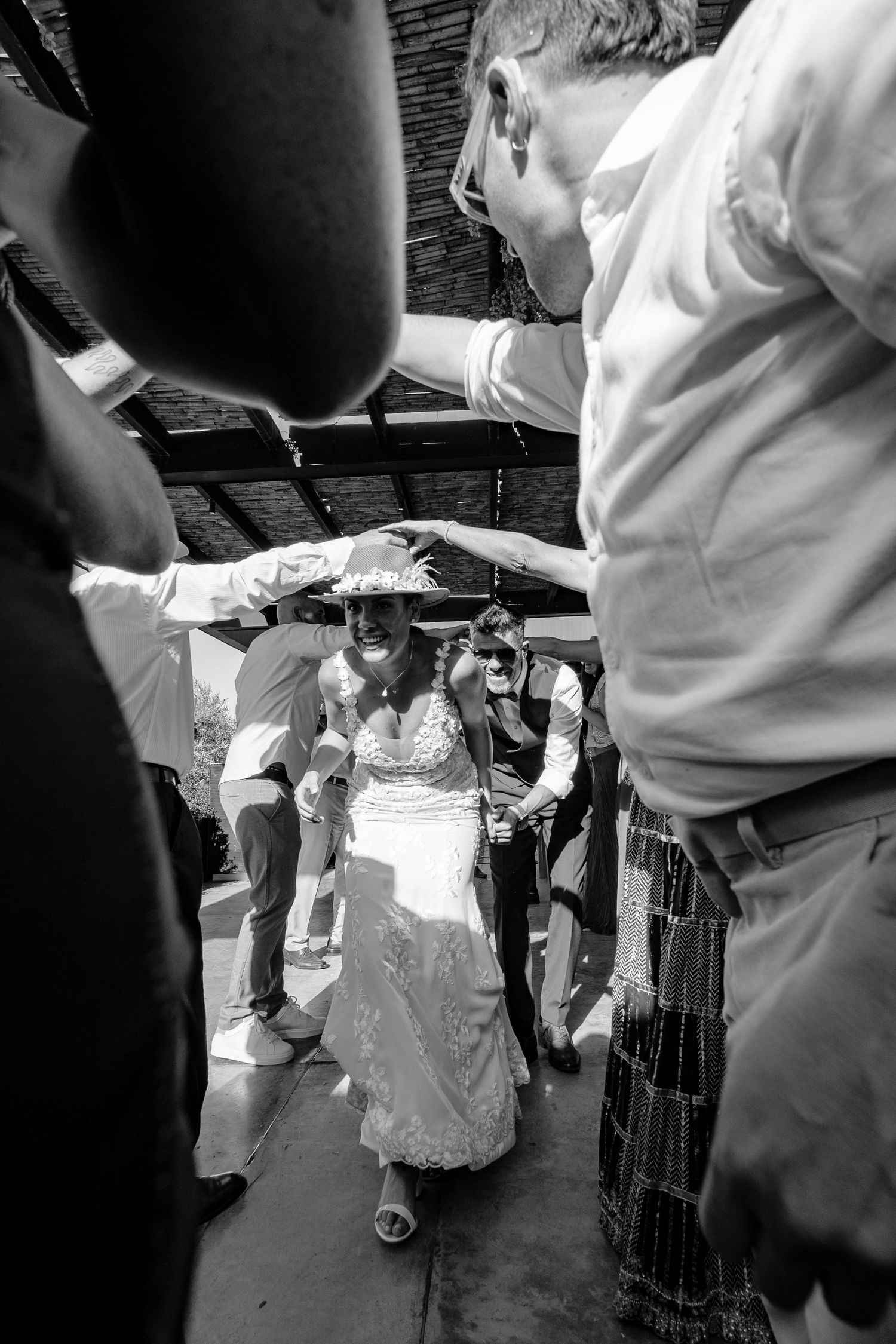 A black and white photo shows a bride in a white dress walking under an archway of raised arms at a celebration.