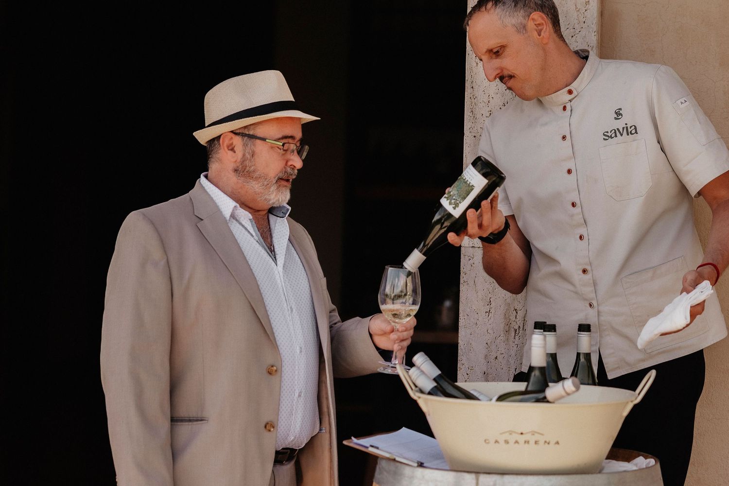 A person in a light colored suit and hat speaks into a microphone while holding a wine glass during an event.