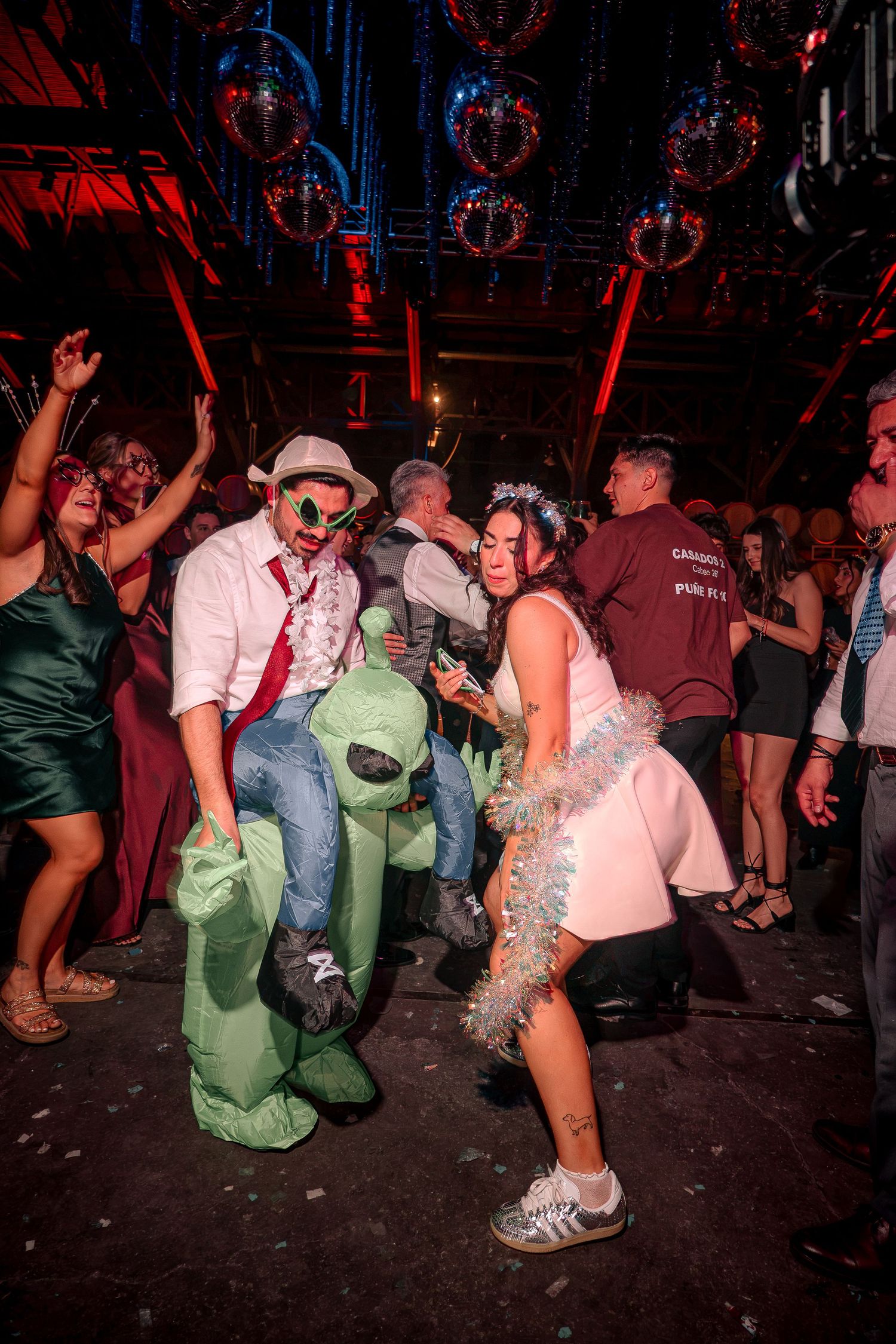 People dancing at a party with disco balls, alien costume, and festive outfits under red lights.