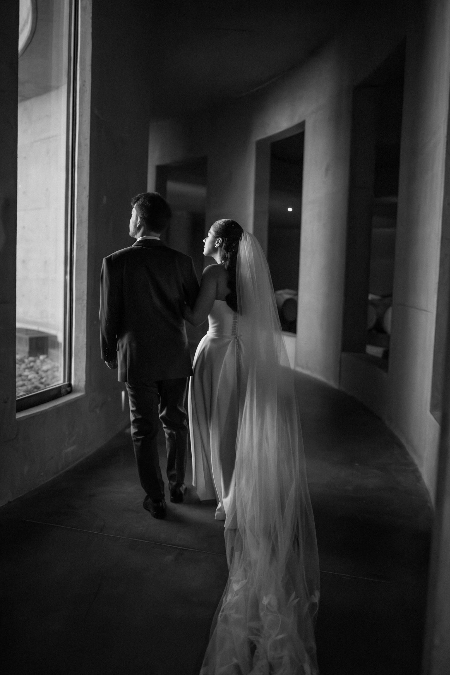 Black and white photo of bride and groom walking down a hallway, bride wearing a long veil trailing behind her. fotógrafos de boda Mendoza
