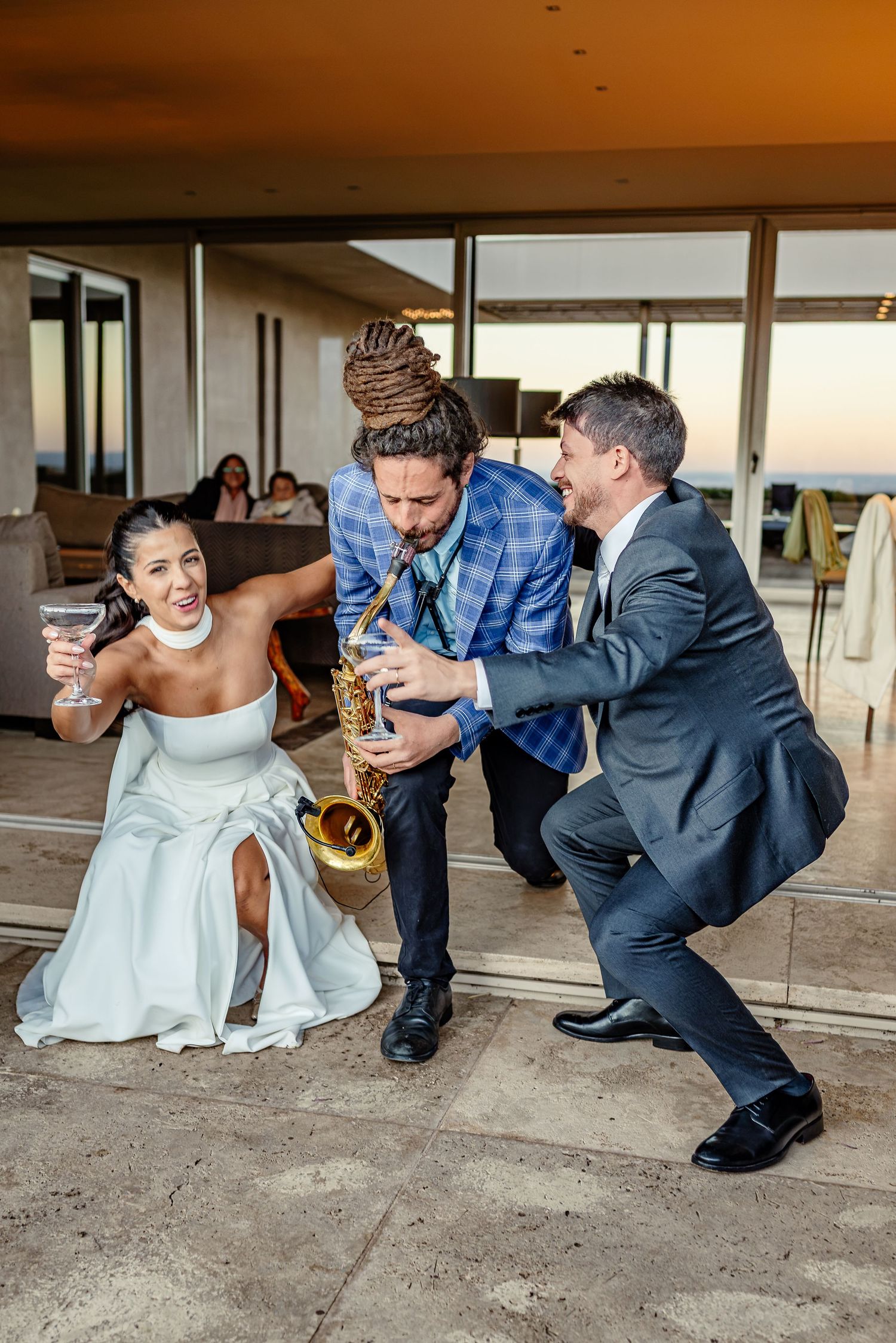 Bride holding cocktail laughs with saxophonist and groomsman dancing at outdoor wedding reception at sunset.
