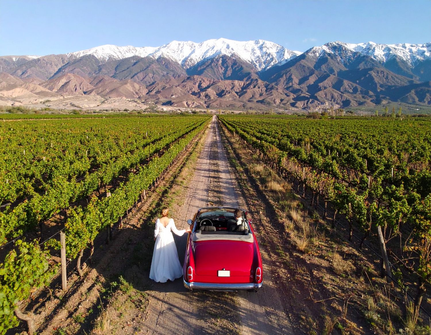 Red vintage car driving through vineyard rows with snow-capped mountains in Mendoza, Argentina.