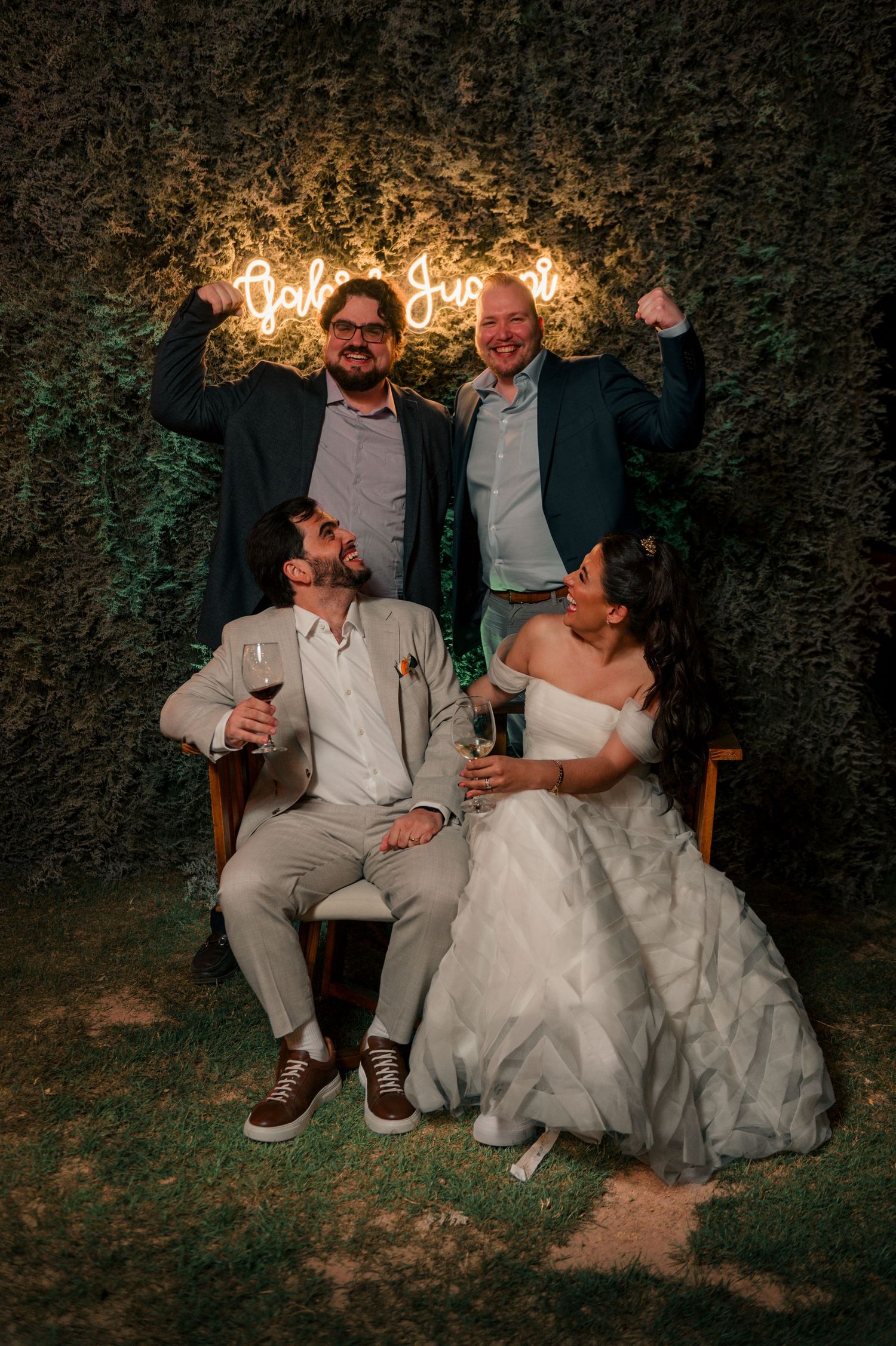 Wedding celebration at night with neon sign glowing against greenery backdrop while couple and guests pose with drinks.