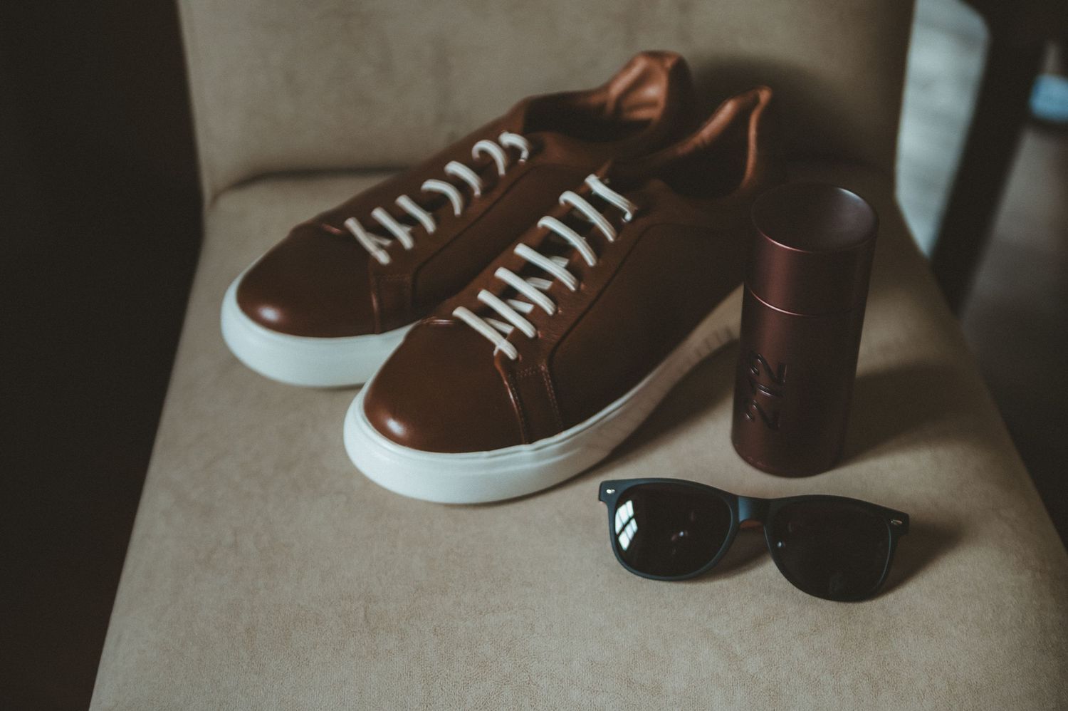 A pair of brown leather sneakers with white soles displayed next to sunglasses and a leather accessory on beige fabric.