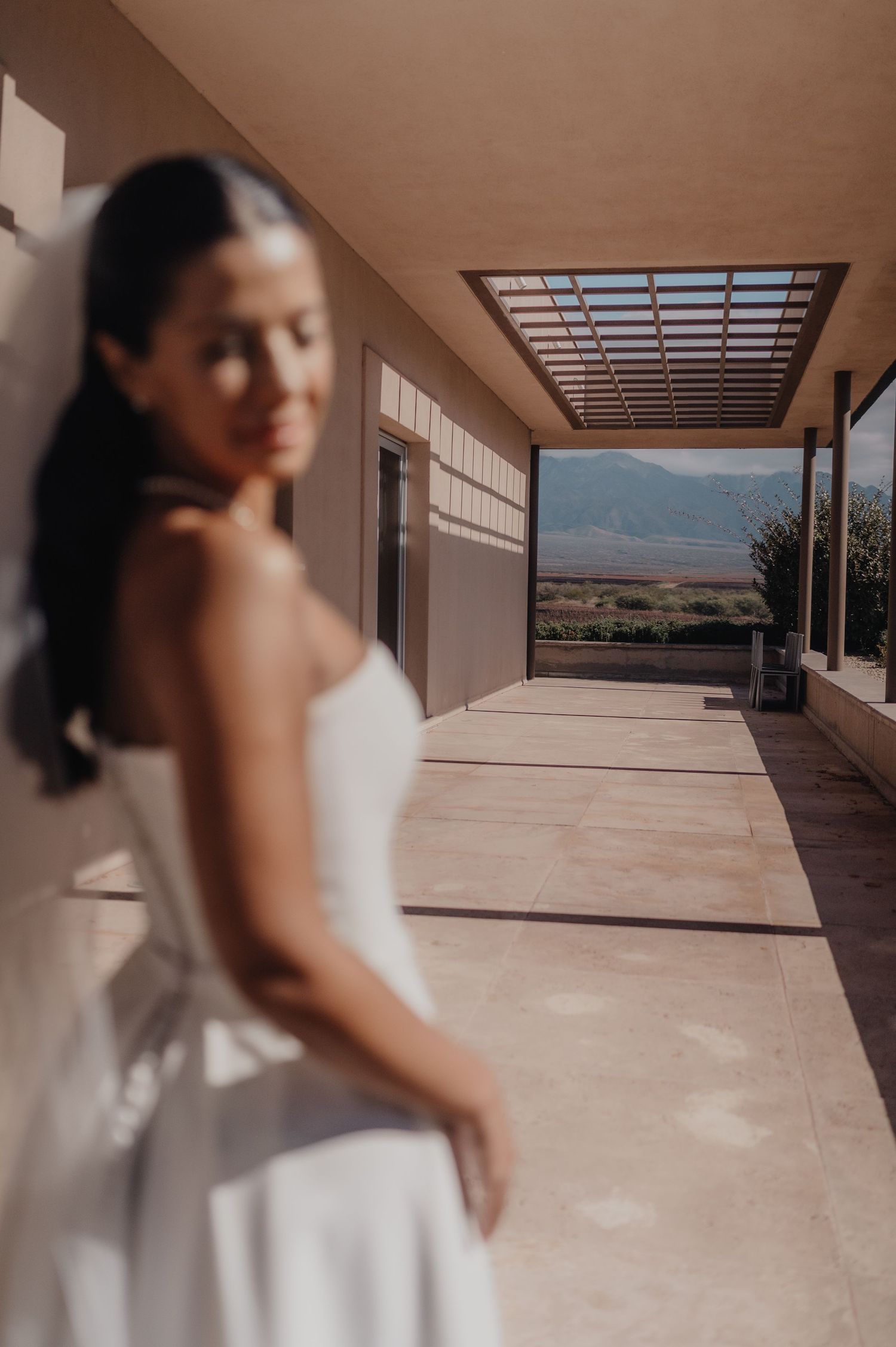 Bride in white strapless gown and veil stands on sunlit terrace with mountain views in background.