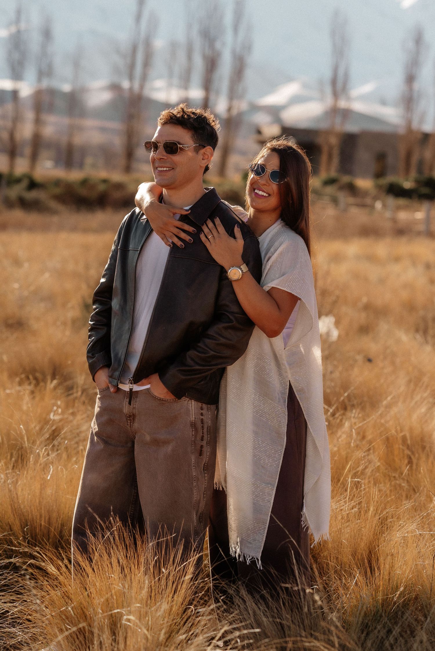 Una pareja con gafas de sol se abraza en un campo dorado, luciendo elegantes atuendos otoñales con montañas de fondo. preboda mendoza