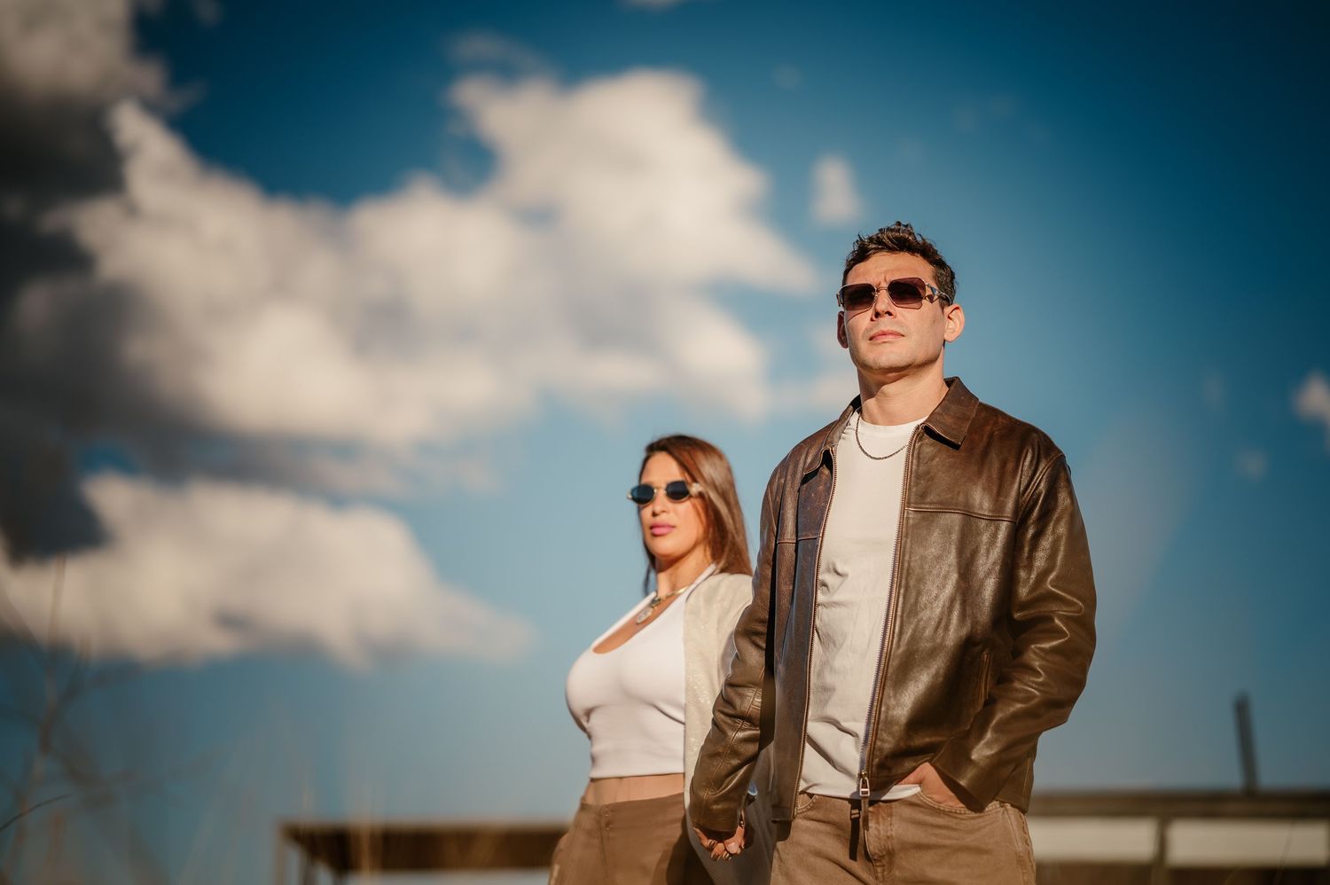 Una pareja elegante, con gafas de sol y chaquetas de cuero marrón, posa al aire libre bajo un cielo azul con nubes blancas. pre boda mendoza